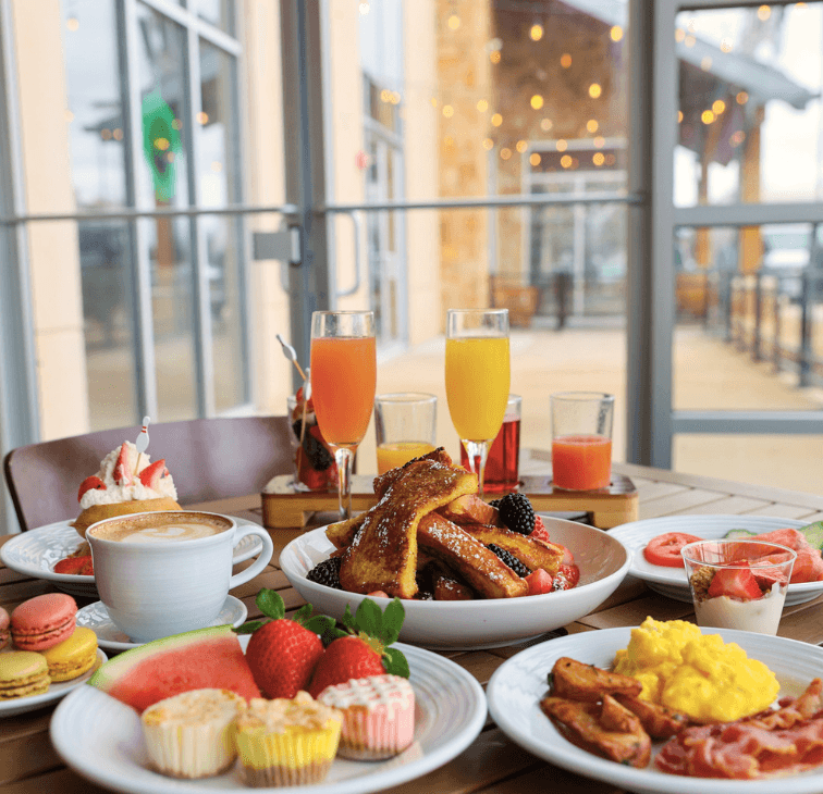 Spread of bottomless brunch items including French toast sticks, macarons, fresh fruit, mimosas, lattes on PNG