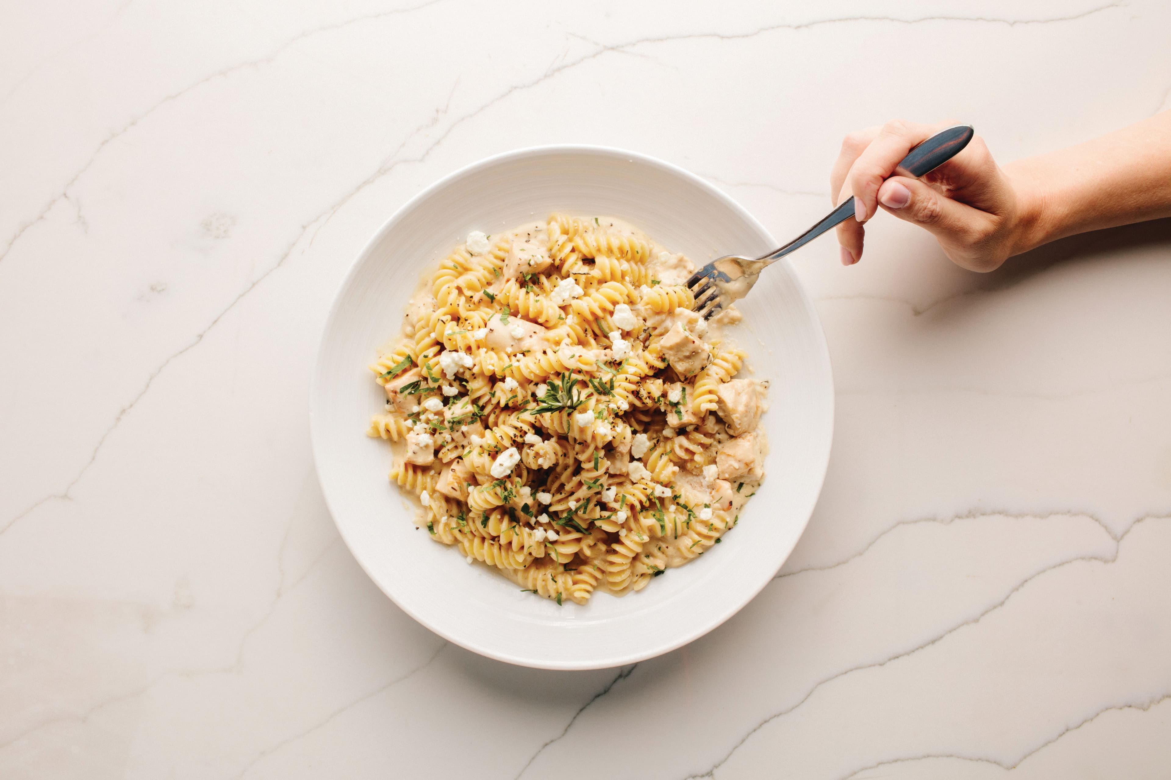 Chicken and goat cheese pasta from Pinstripes on a white marble background, with a hand grabbing some pasta with a fork.