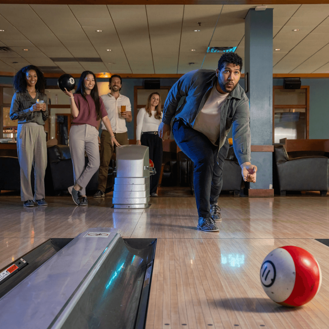 Group of friends laughing and holding bowling balls during a lively game at Pinstripes, promoting the Prime Time Play Unlimited Bowling Pass
