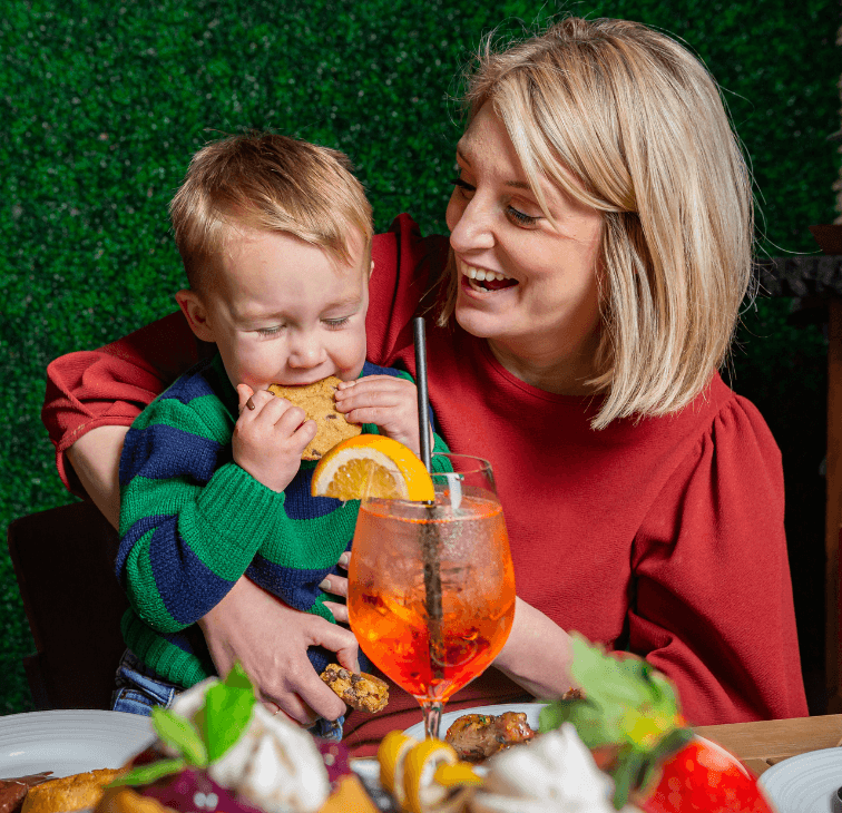Mom laughing while holding her small son biting into a cookie at Pinstripes all-you-can-eat brunch