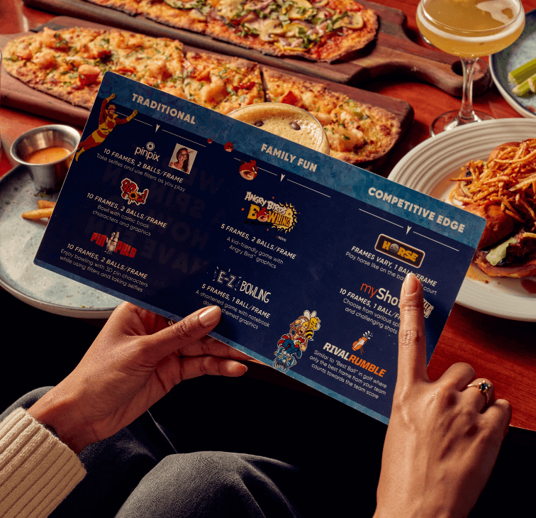 Person holding a Pinstripes gaming card showing bowling game options, with food and drinks in the background