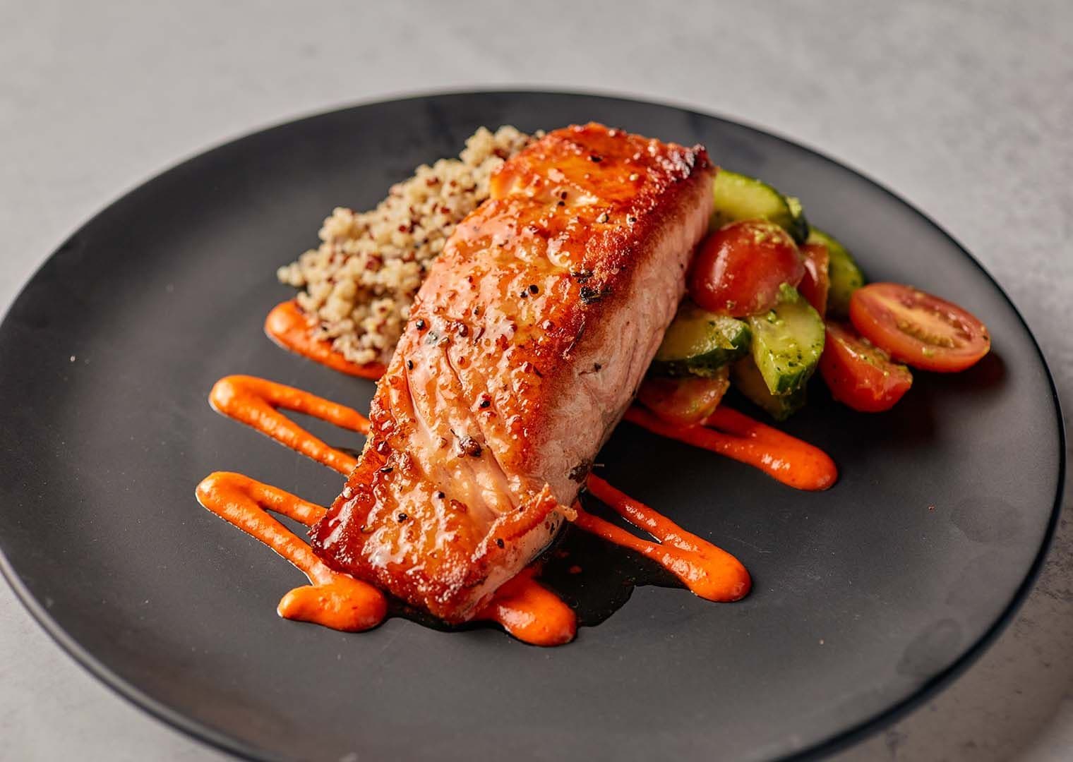 Maple Glazed Salmon from Pinstripes with hand‑cut herb‑marinated salmon, Mediterranean grains, cucumber, tomato, avocado, and pesto on a plate.