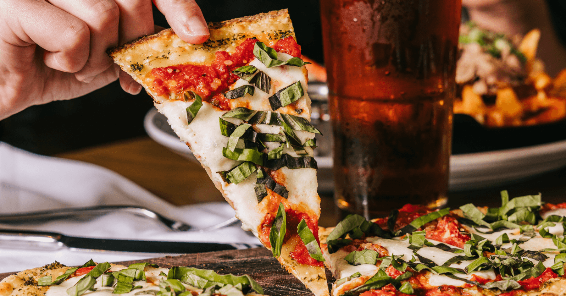 Hand holding a slice of Margherita Flatbread with a glass of beer in the background.