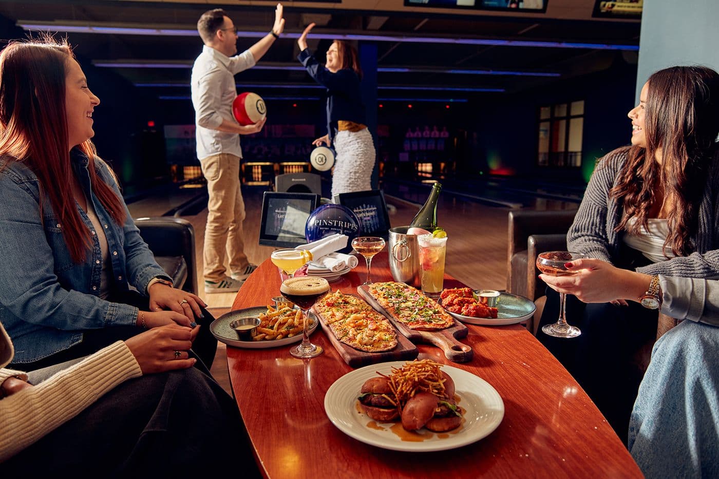 Friends enjoying themselves on the bowling lanes at Pinstripes, with some eating and others high‑fiving on the lanes.