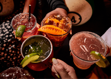 Hands holding a mule, margarita, and old fashioned with Pinstripes logo on orange peel, bowling balls in background