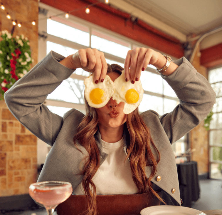 Woman at brunch holding two sunny-side-up eggs in front of her eyes in a playful pose