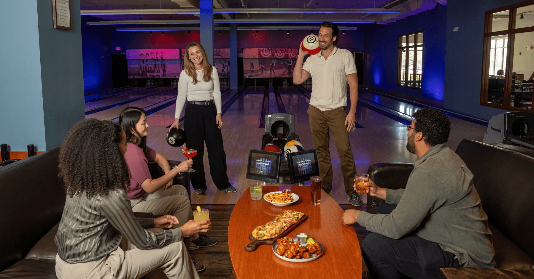 Five friends at a bowling lane with two holding bowling balls and others enjoying food and cocktails on lounge seating.