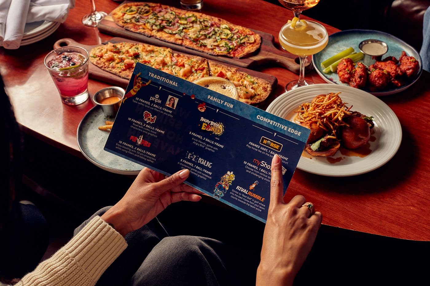 Hand holding a Pinstripes bowling games card in front of a table loaded with shareable bites, showing the game options while guests enjoy appetizers and flatbreads.