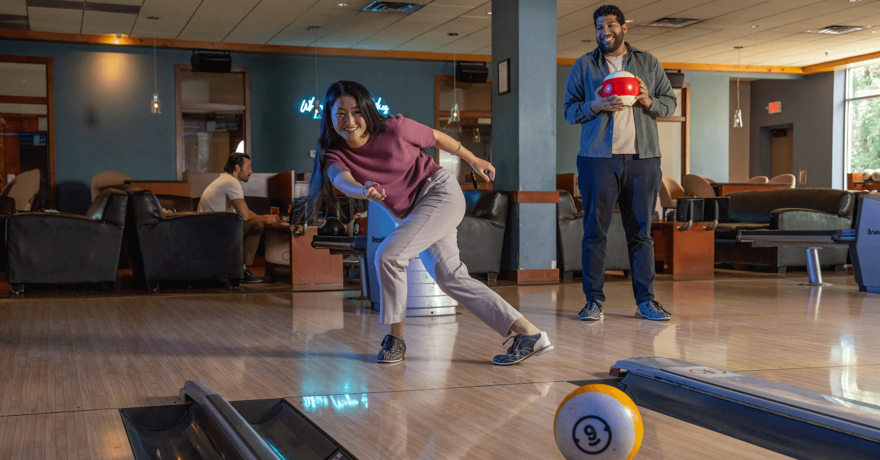 Woman rolling a bowling ball down the lane while a friend watches from behind.