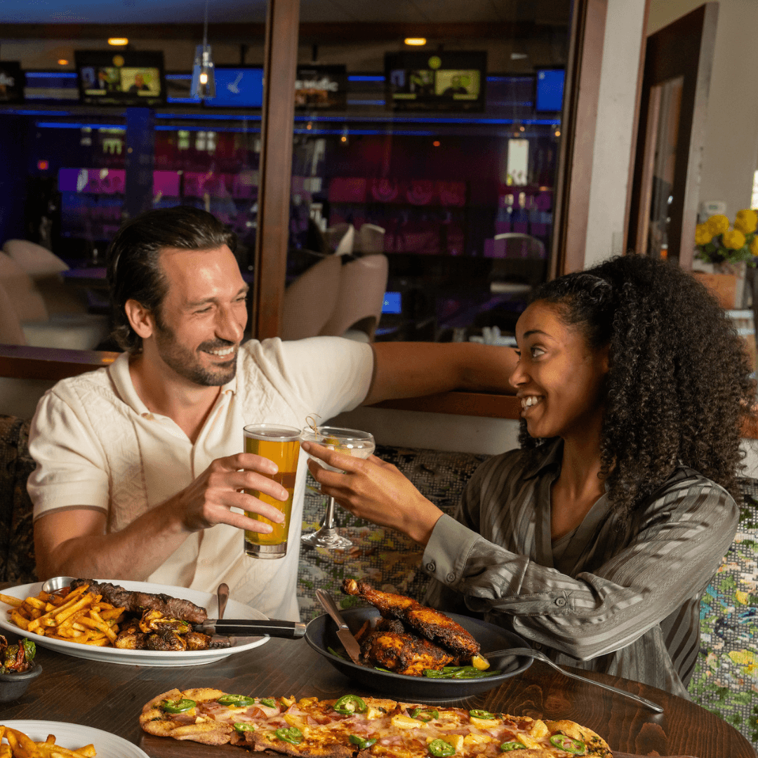 Man and woman raising glasses over a table with Chimi Steak, Hawaiian Flatbread, Rustic Chicken and shared dishes.